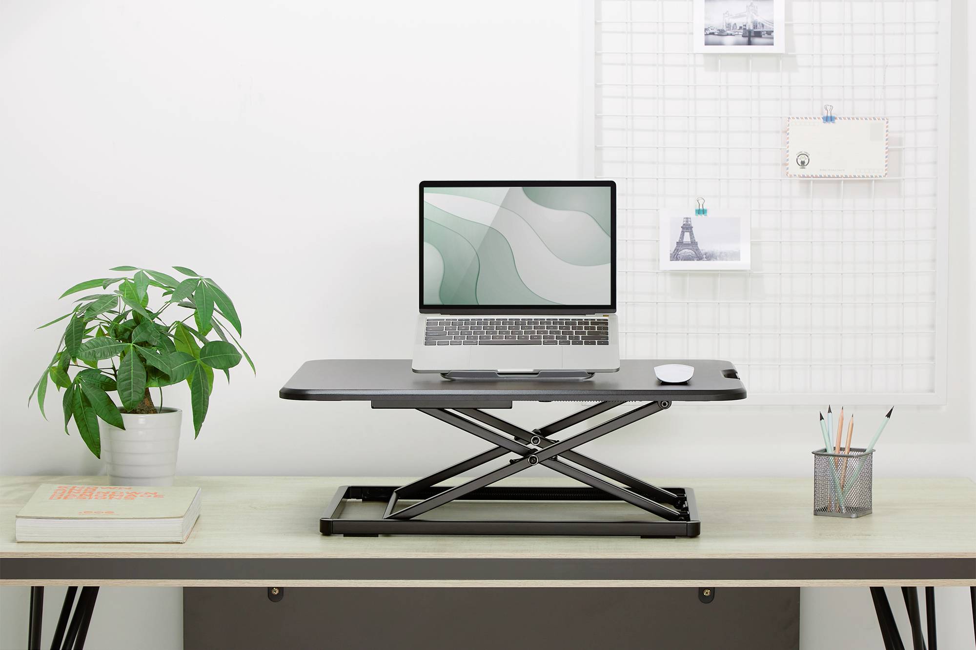 Ein höhenverstellbarer Schreibtischkonverter mit Laptop darauf, umgeben von Büropflanze, Notizen an einem Gitter und Schreibutensilien.