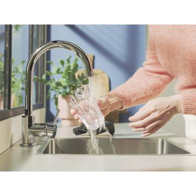 Eine Person spült ein Glas unter fließendem Wasser in der Küche. Im Hintergrund sind Pflanzen und Küchenutensilien sichtbar.