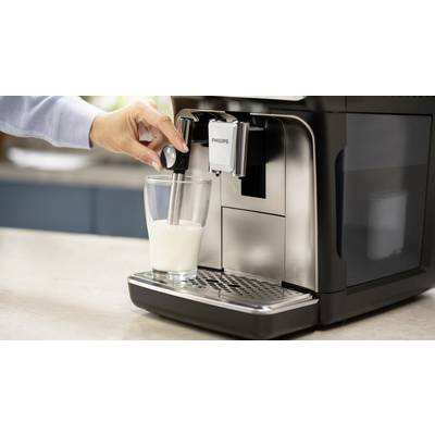 Hand hält einen Glas unter einem Milchaufschäumer an einer Kaffeemaschine. Milch wird aufgeschäumt. Küche im Hintergrund erkennbar.
