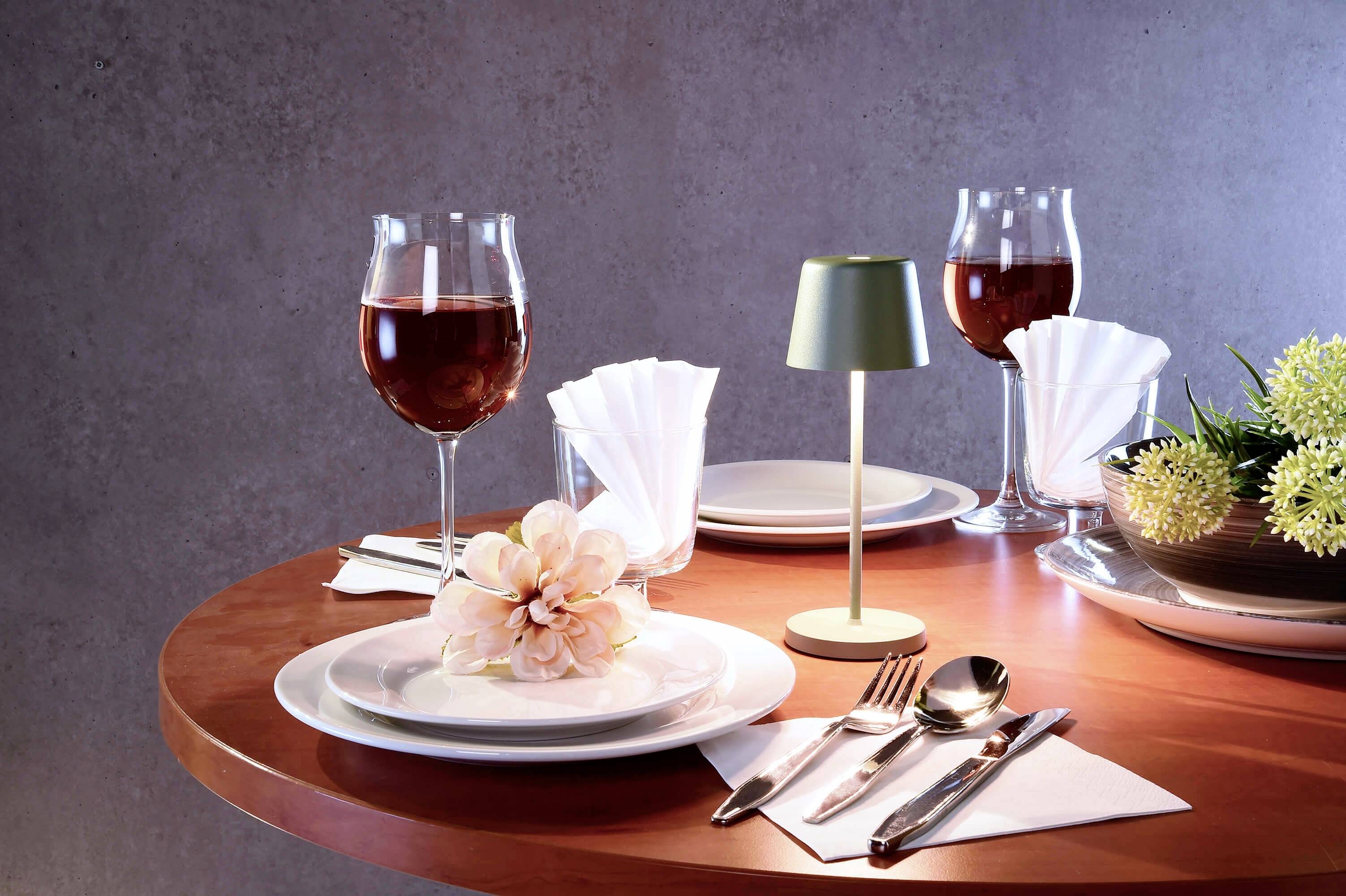 Ein runder Tisch ist elegant gedeckt mit Weingläsern, Besteck, Servietten und Blumen. Zwei Gläser Rotwein stehen bereit.