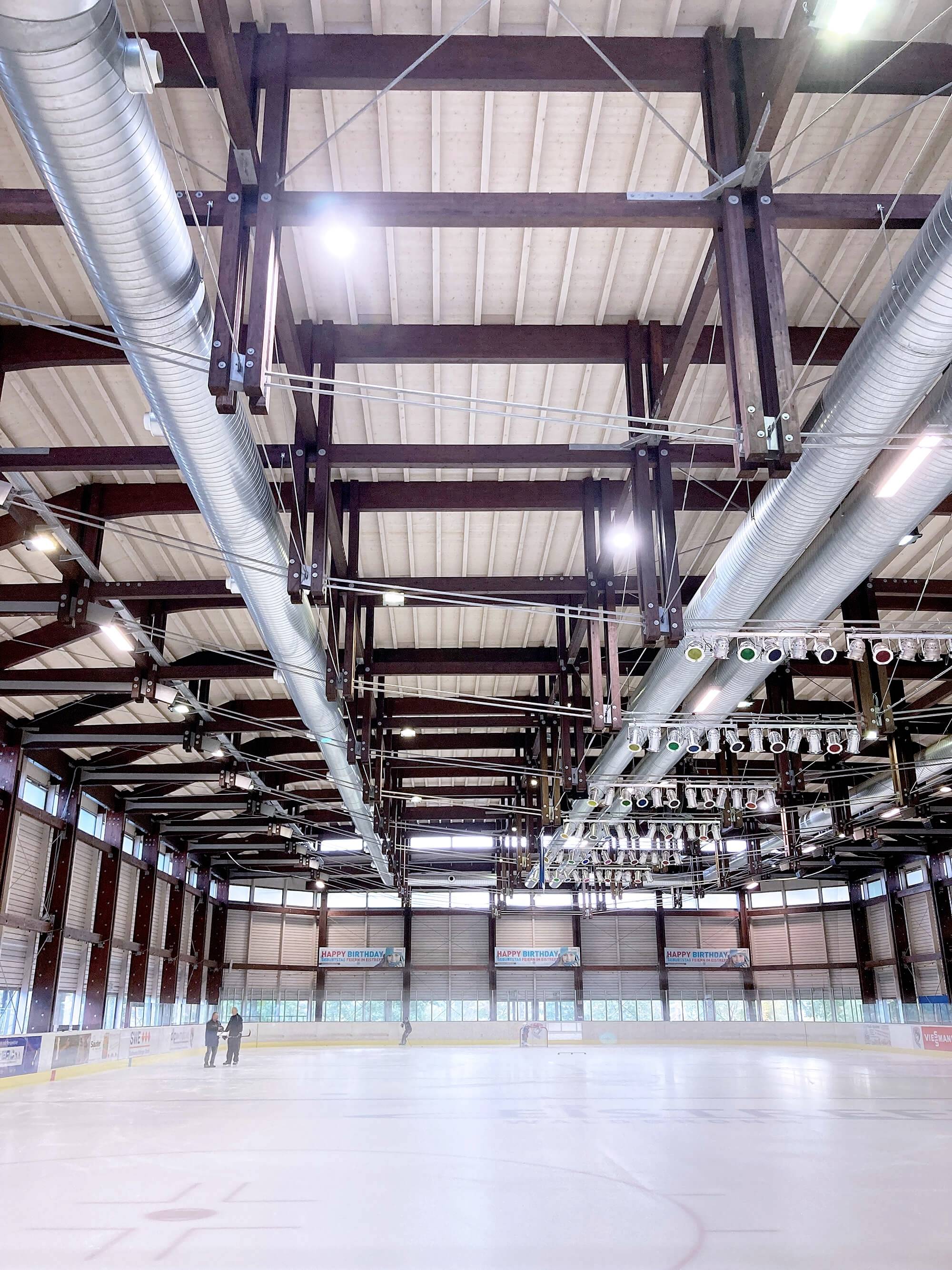 Eisbahn in einer Halle; leere Spielfläche mit sichtbarer Holzdachkonstruktion und Lüftungsrohren; zwei Personen am Rand.