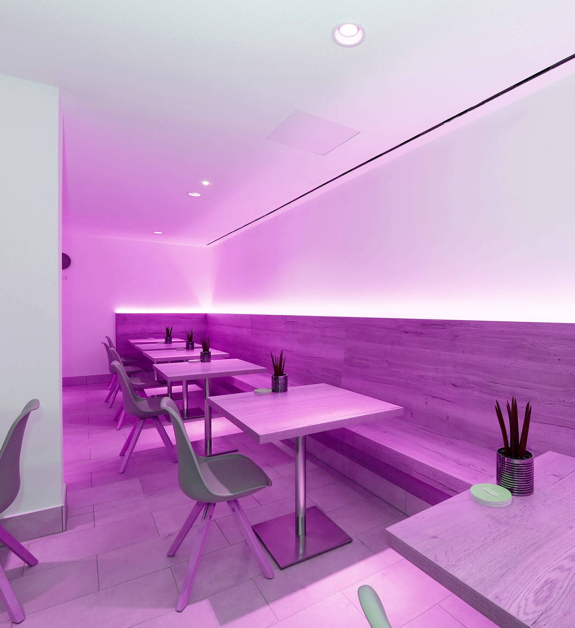 Ein modernes Café mit minimalistischem Design, rosa Beleuchtung, Holztischen, grauen Stühlen und kleinen Pflanzen als Dekoration.
