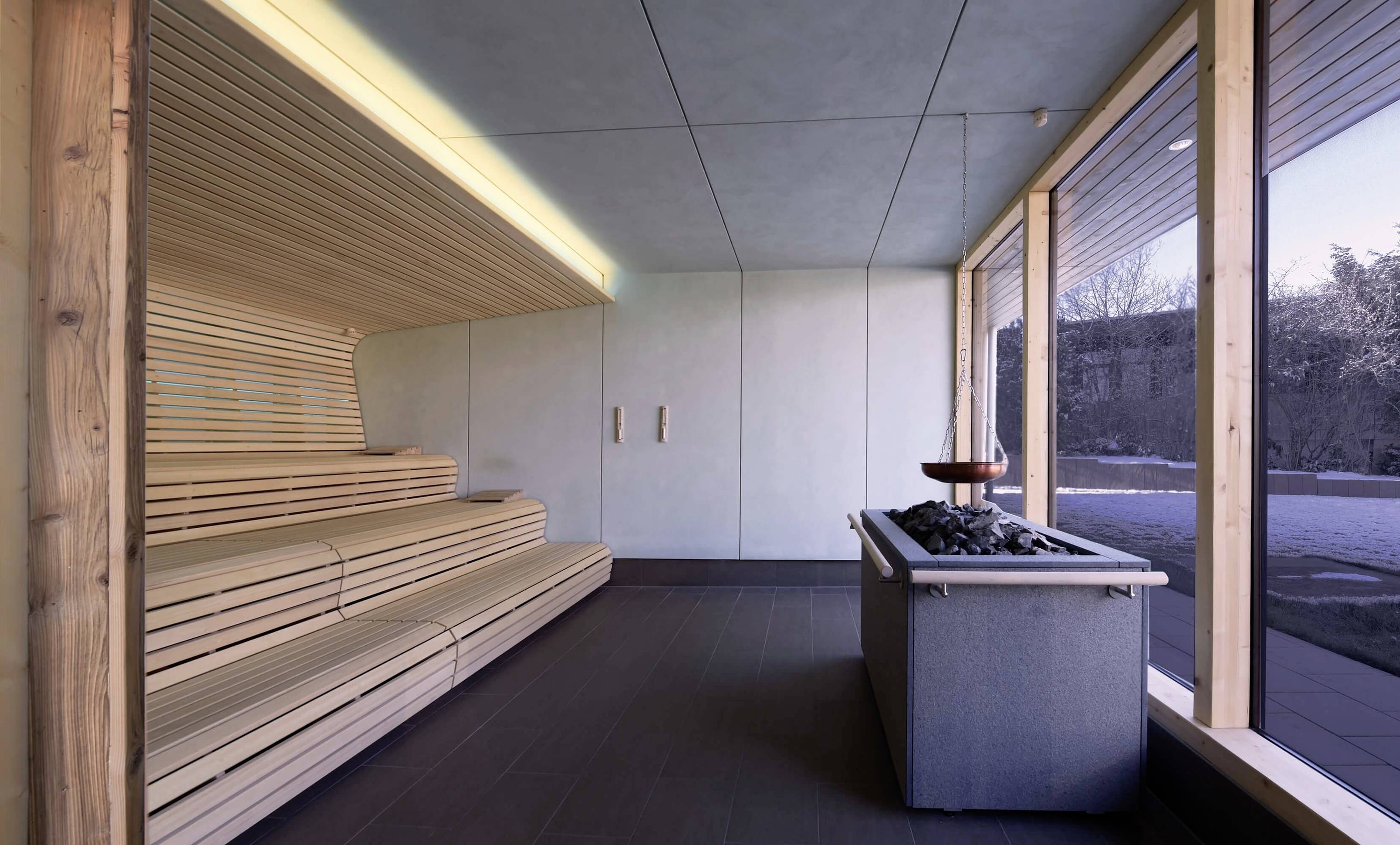 Eine moderne Sauna mit gestuften Holzbänken, einem Saunaofen im Vordergrund und großen Fenstern mit Blick auf eine winterliche Landschaft.