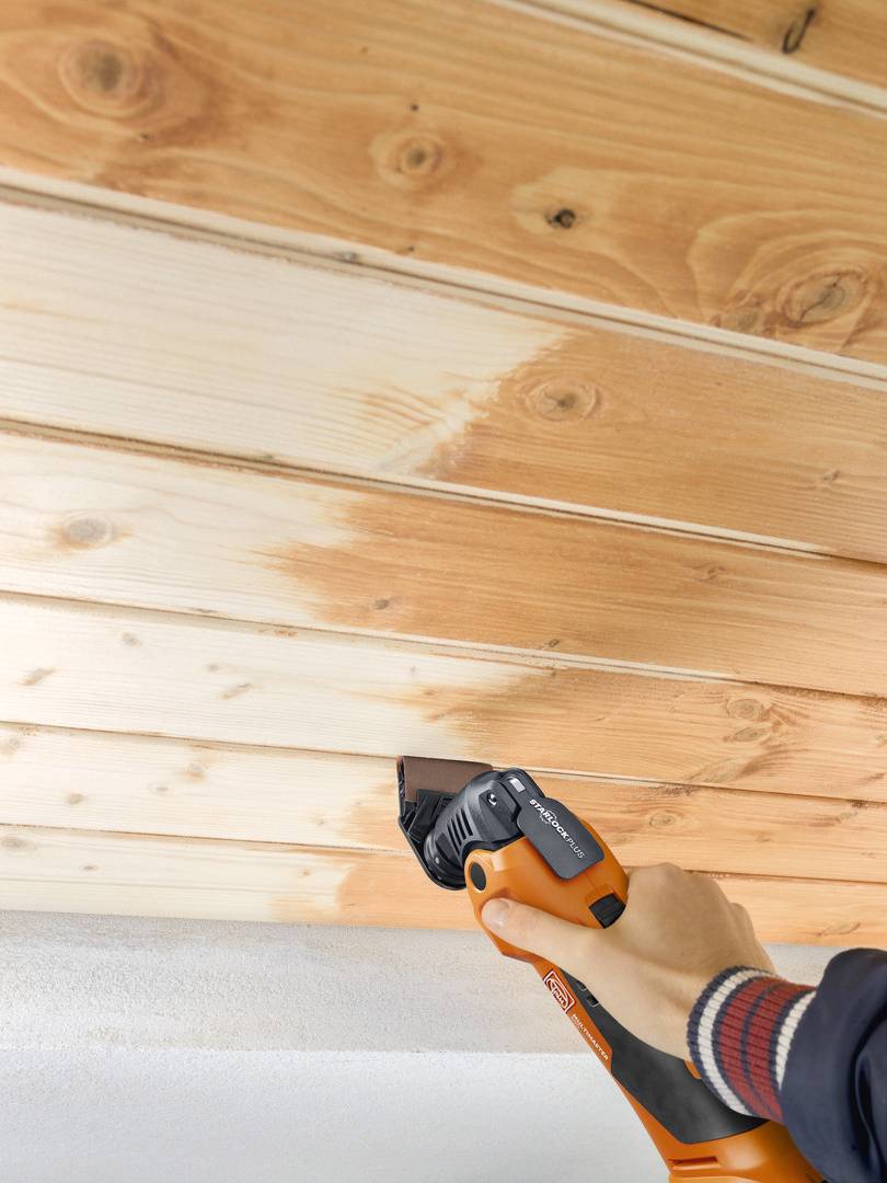 Eine Hand hält ein orangefarbenes Elektrowerkzeug, das Holzpaneele an einer Decke schleift. Das Holz zeigt teilweise abgeschliffene Bereiche.