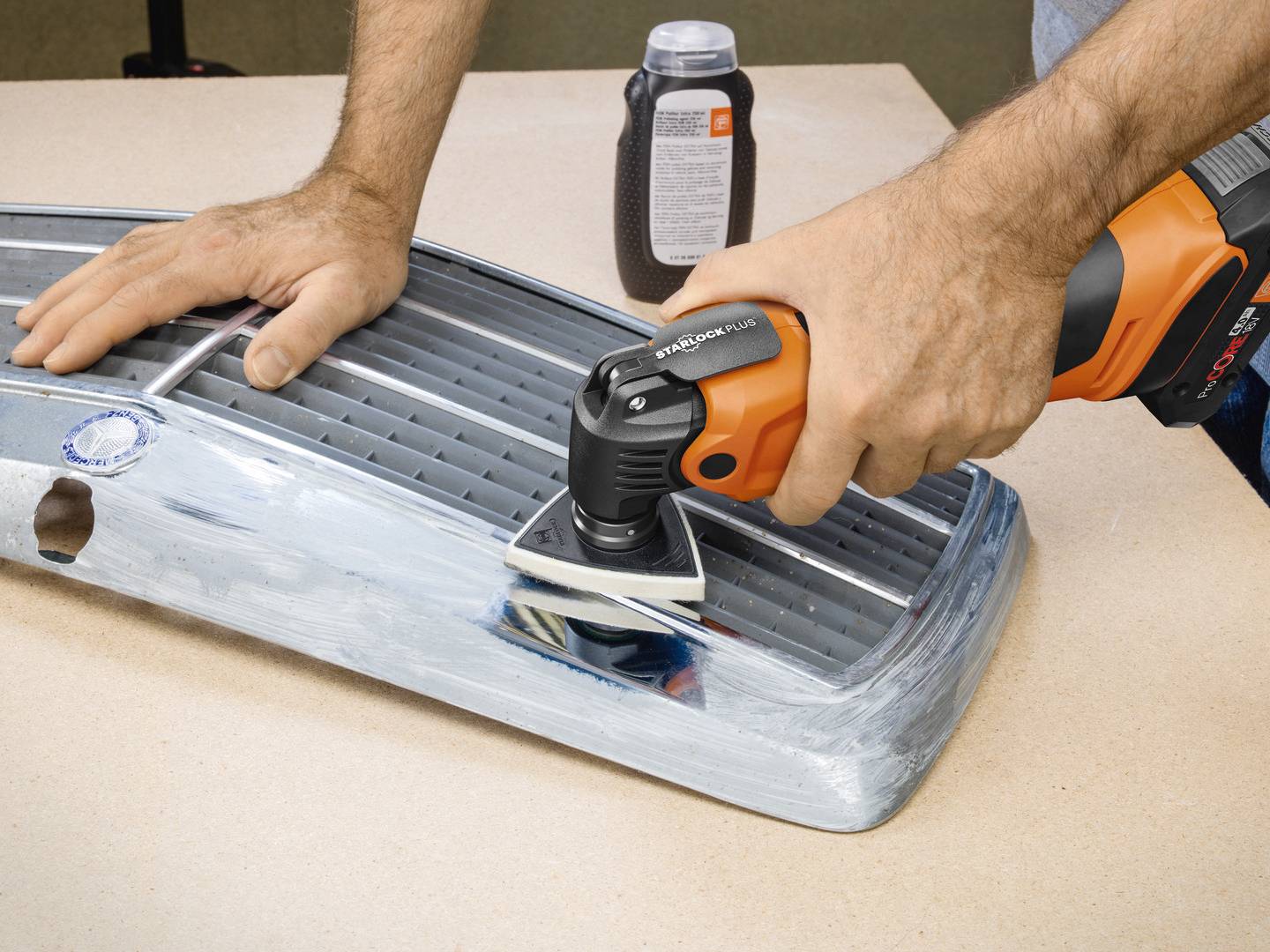 Person schleift einen Autostoßfänger mit einem orange-schwarzen Elektrowerkzeug. Ein Poliermittelbehälter steht im Hintergrund.
