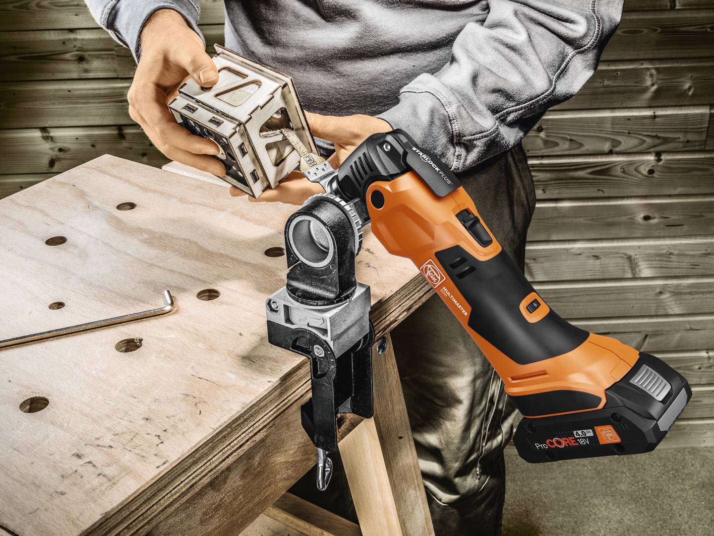 Eine Person verwendet ein orange-schwarzes Multifunktionswerkzeug, um einen Holzblock in einem Schraubstock auf einem Arbeitstisch zu bearbeiten.