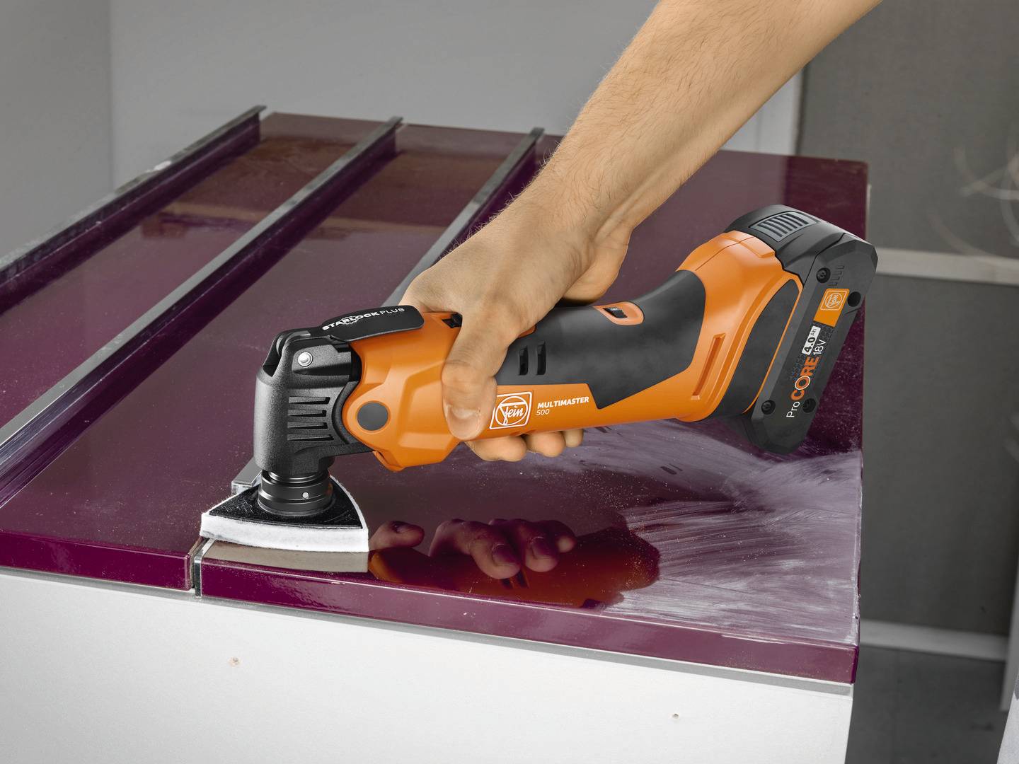 Eine Person verwendet ein oranges Multifunktionswerkzeug, um Lack von einer Oberfläche zu entfernen.