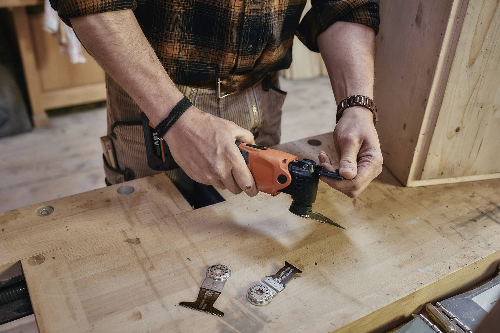 Eine Person arbeitet an einem Holztisch mit einem oszillierenden Werkzeug. Zwei Ersatzklingen liegen vor ihnen auf dem Tisch.