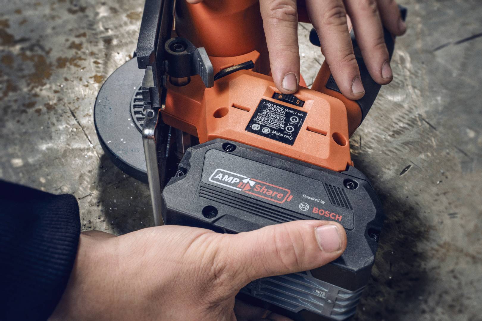 Zwei Hände befestigen einen Akku an einem orangefarbenen Elektrogerät. Das Gerät liegt auf einer Metalloberfläche.