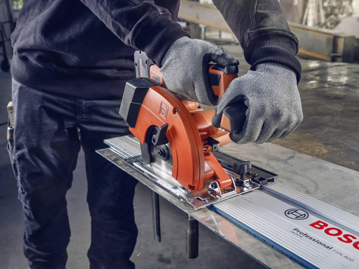 Eine Person sägt mit einer orangen Handkreissäge durch ein Metallstück auf einer Werkbank. Schutzkleidung inklusive Handschuhe wird getragen.