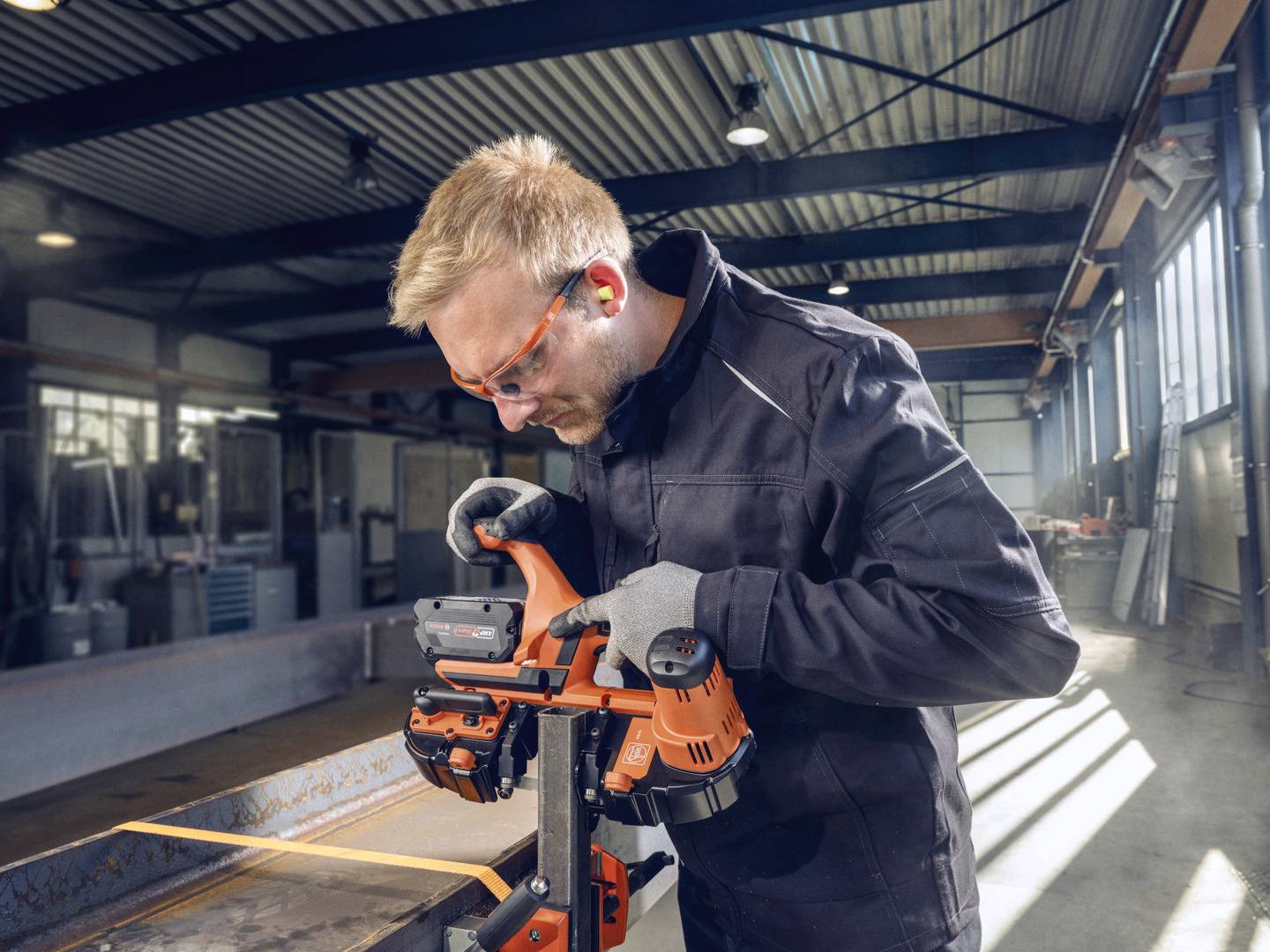 Ein Mann in Arbeitskleidung benutzt ein orangefarbenes Elektrowerkzeug in einer Werkstatt. Er trägt Schutzbrille und Arbeitshandschuhe.