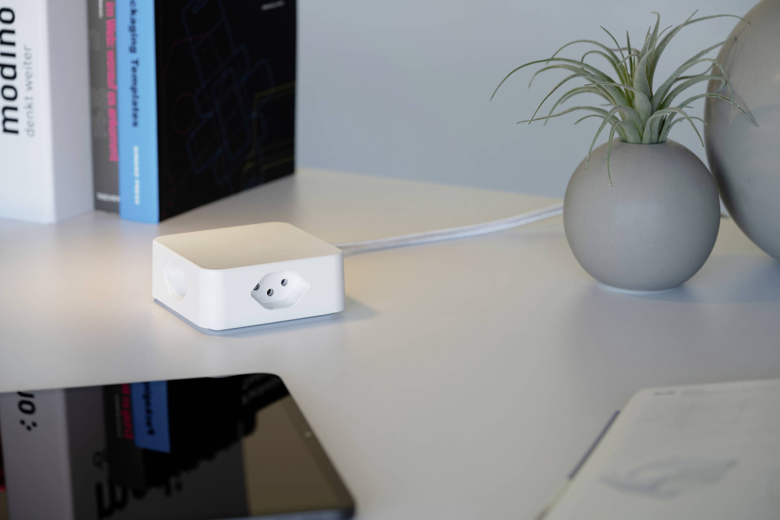 Ein weißes elektronisches Gerät mit Anschluss liegt auf einem Tisch neben einer Vase mit Pflanze und einem schwarzen Tablet.