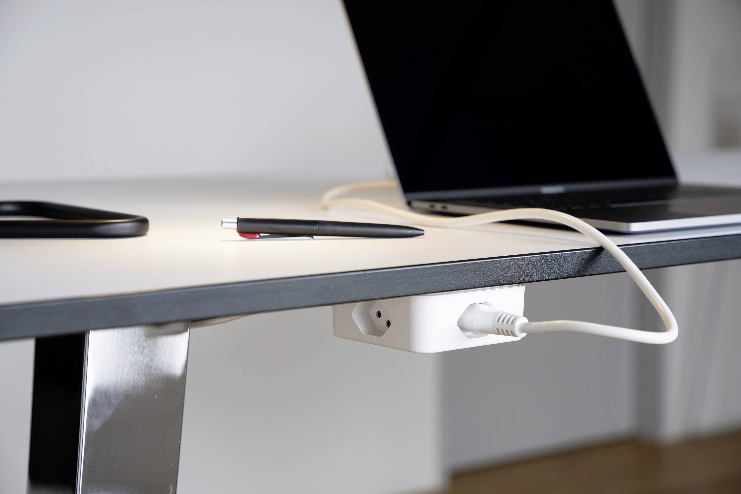 Laptop auf Schreibtisch, mit angeschlossenem Kabel. Ein Stift liegt daneben. Arbeitsumgebung in modernem Bürodesign.