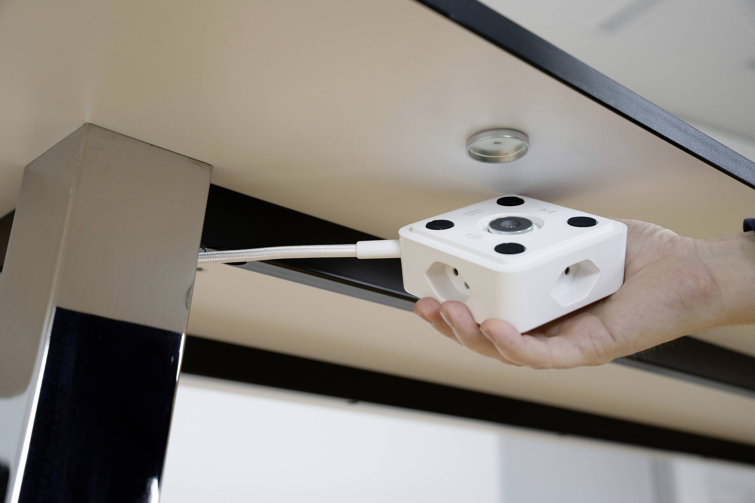 Eine Hand hält einen kompakten Sensor mit Kabelanschluss unter einen Tisch. Der Sensor ist weiß mit schwarzen Punkten an den Ecken.
