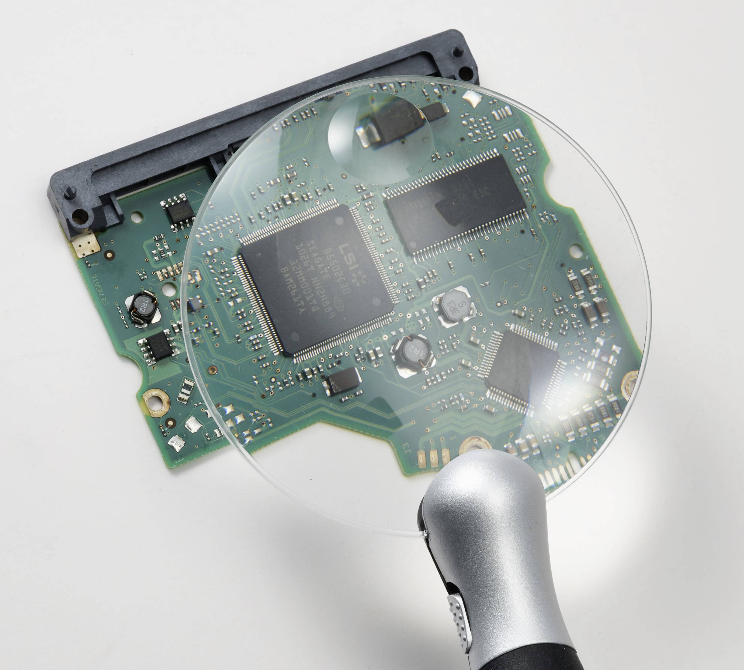 Vergrößerte Ansicht einer grünen Leiterplatte mit vielen kleinen elektrischen Komponenten. Eine Lupe fokussiert auf die Platine.