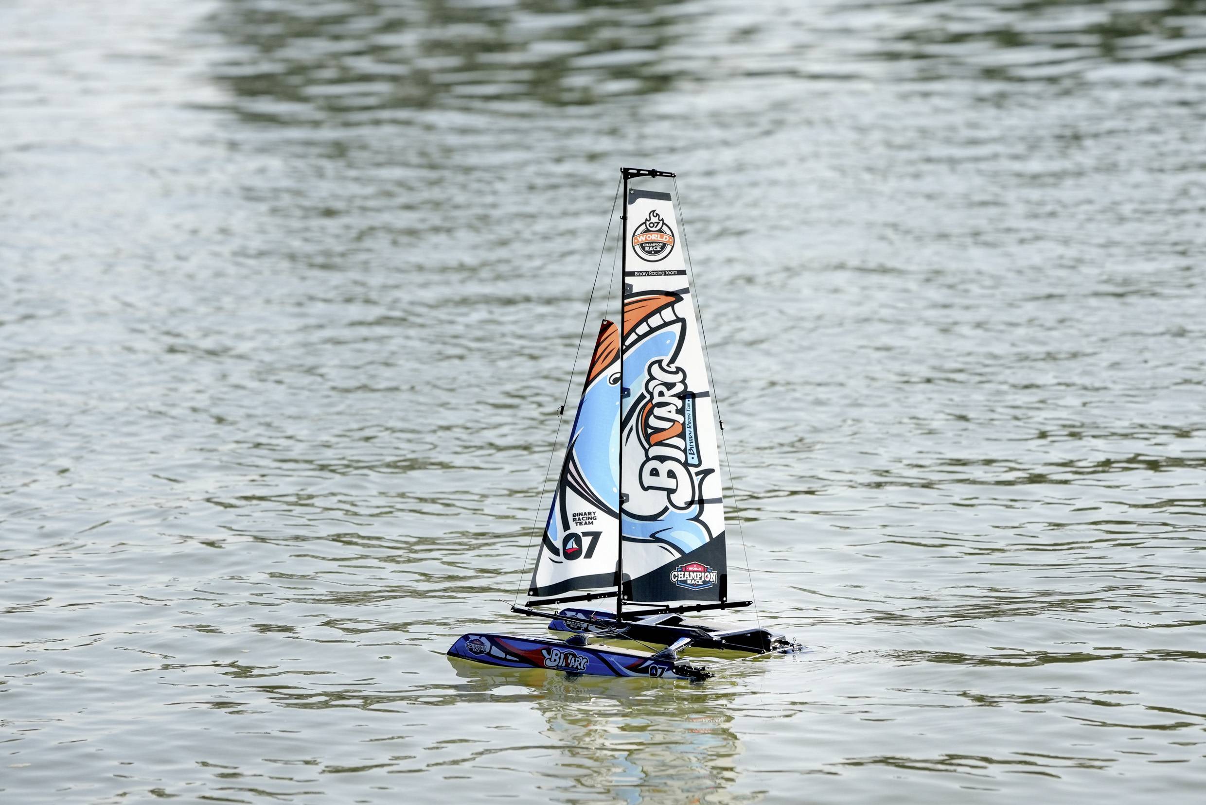 Ein kleines ferngesteuertes Segelboot mit buntem Segel fährt auf einem See. Das Boot hat die Nummer '07' und bewegt sich durch ruhiges Wasser.