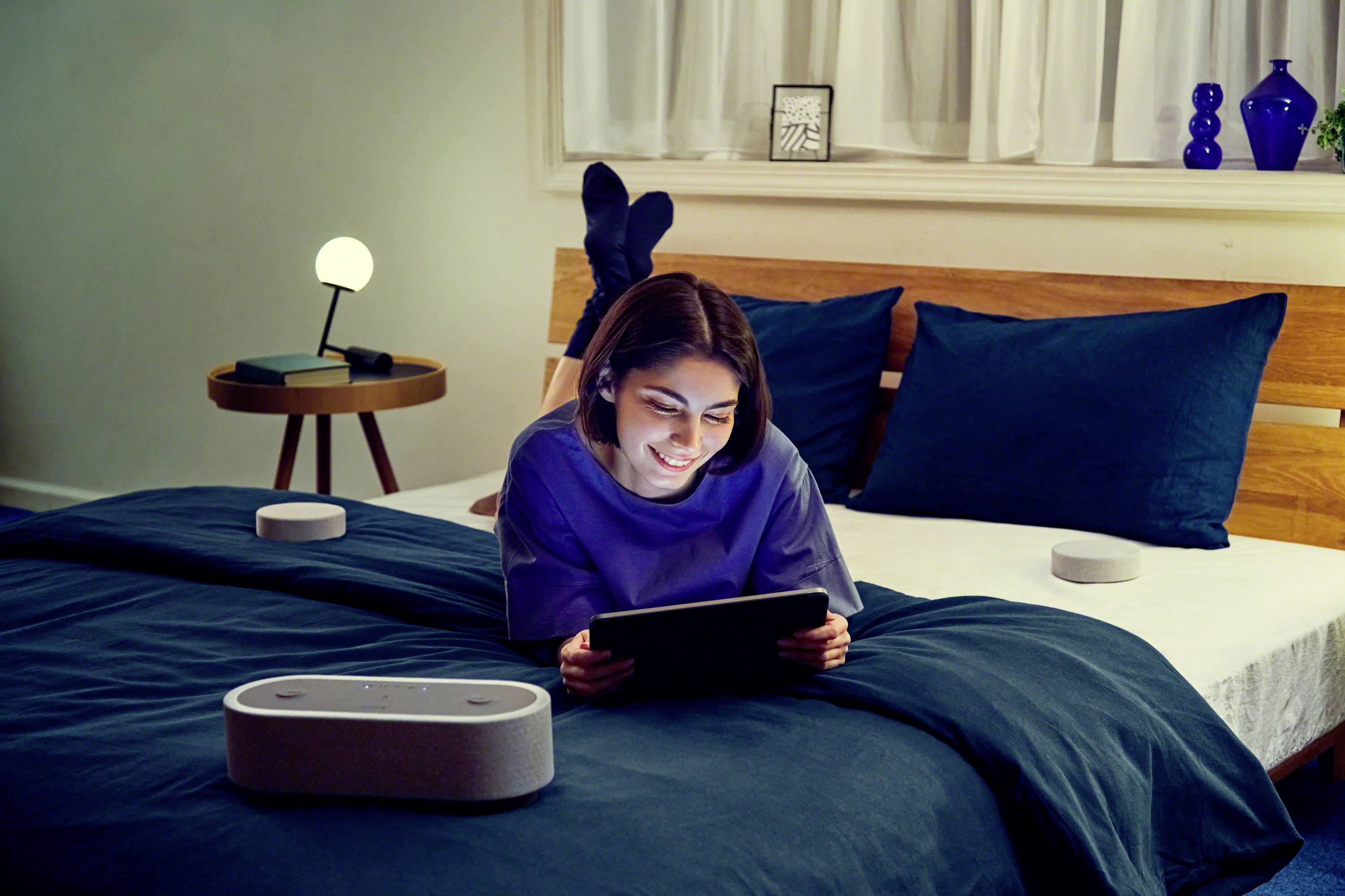 Eine Frau liegt entspannt auf einem Bett und schaut lächelnd auf ein Tablet. Smarte Lautsprecher sind um sie herum platziert.