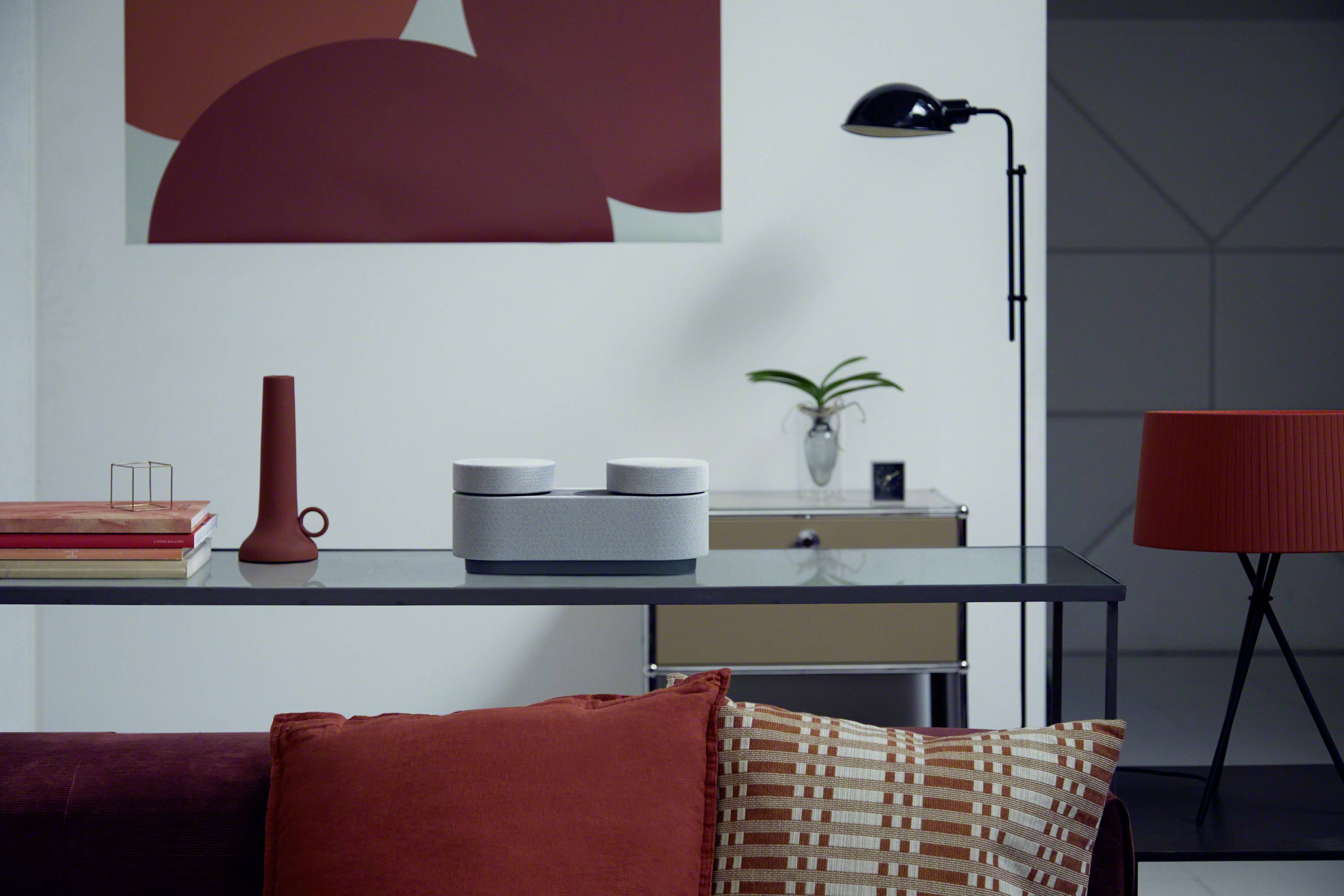 Ein moderner Raum mit einem Glastisch, darauf Deko-Objekte und Lautsprecher. Rote und graue Akzente dominieren das Design. Im Hintergrund ein abstraktes Kunstwerk.