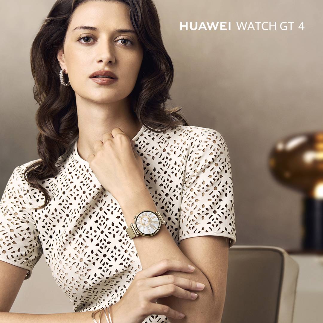 Eine Frau trägt eine weiße, durchbrochene Bluse und eine Huawei Watch GT 4. Sie sitzt auf einem Stuhl. Links oben das Huawei-Logo.