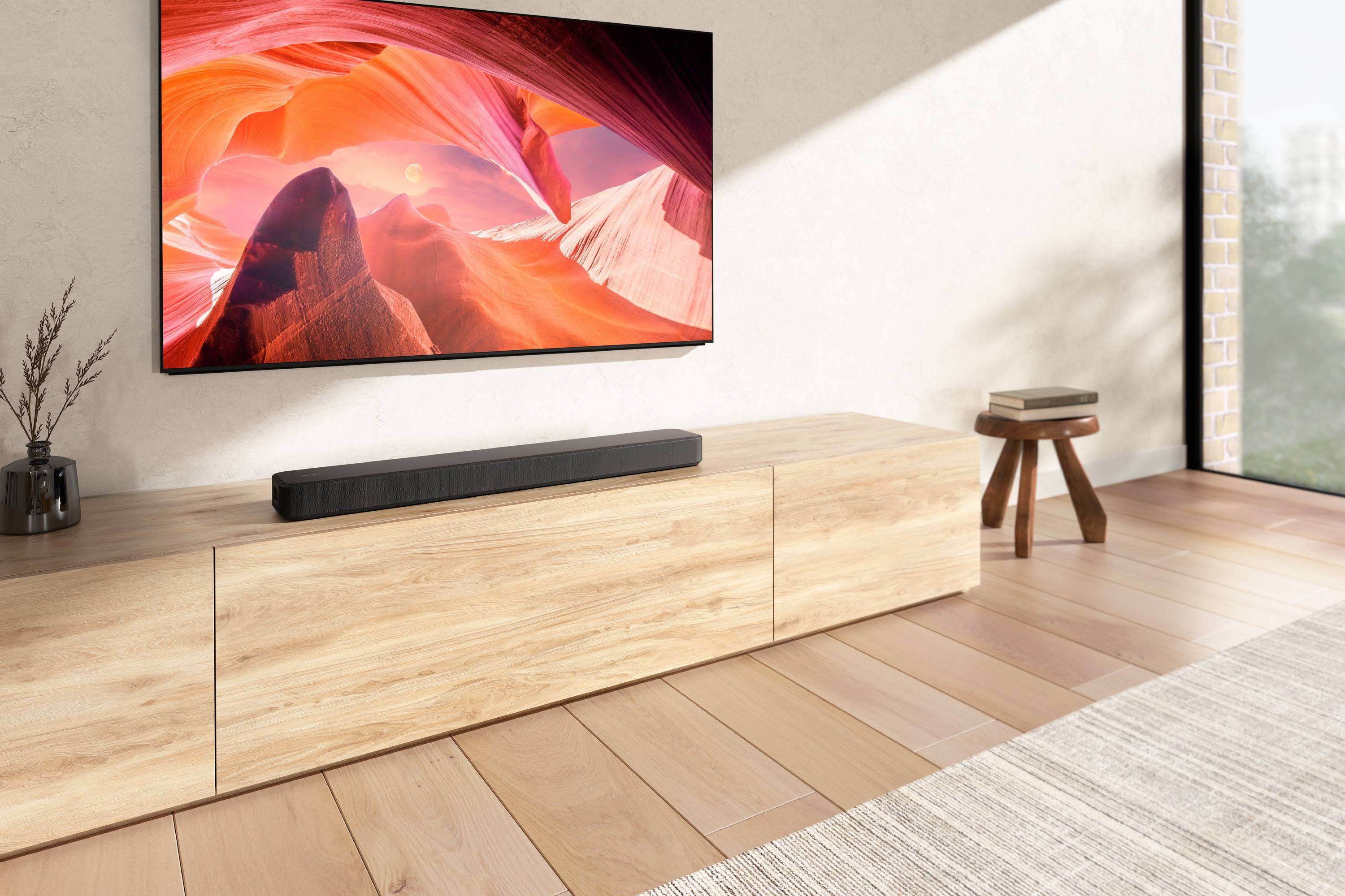 Ein modernes Wohnzimmer mit hellem Holzboden. Ein großer, farbenfroher Fernseher hängt an der Wand über einem Holzschrank.