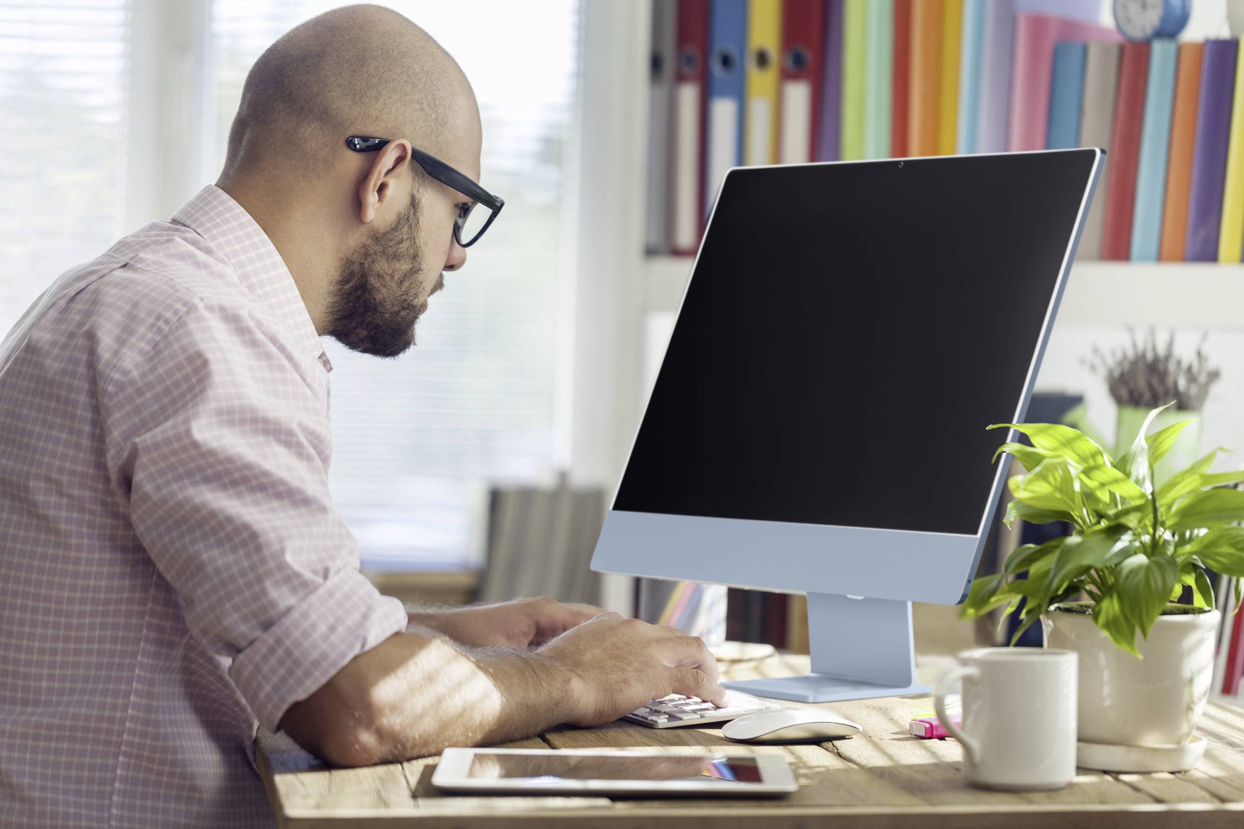 Ein Mann mit Brille arbeitet an einem Computertisch. Neben ihm steht eine Pflanze, und im Hintergrund sind Aktenordner zu sehen.