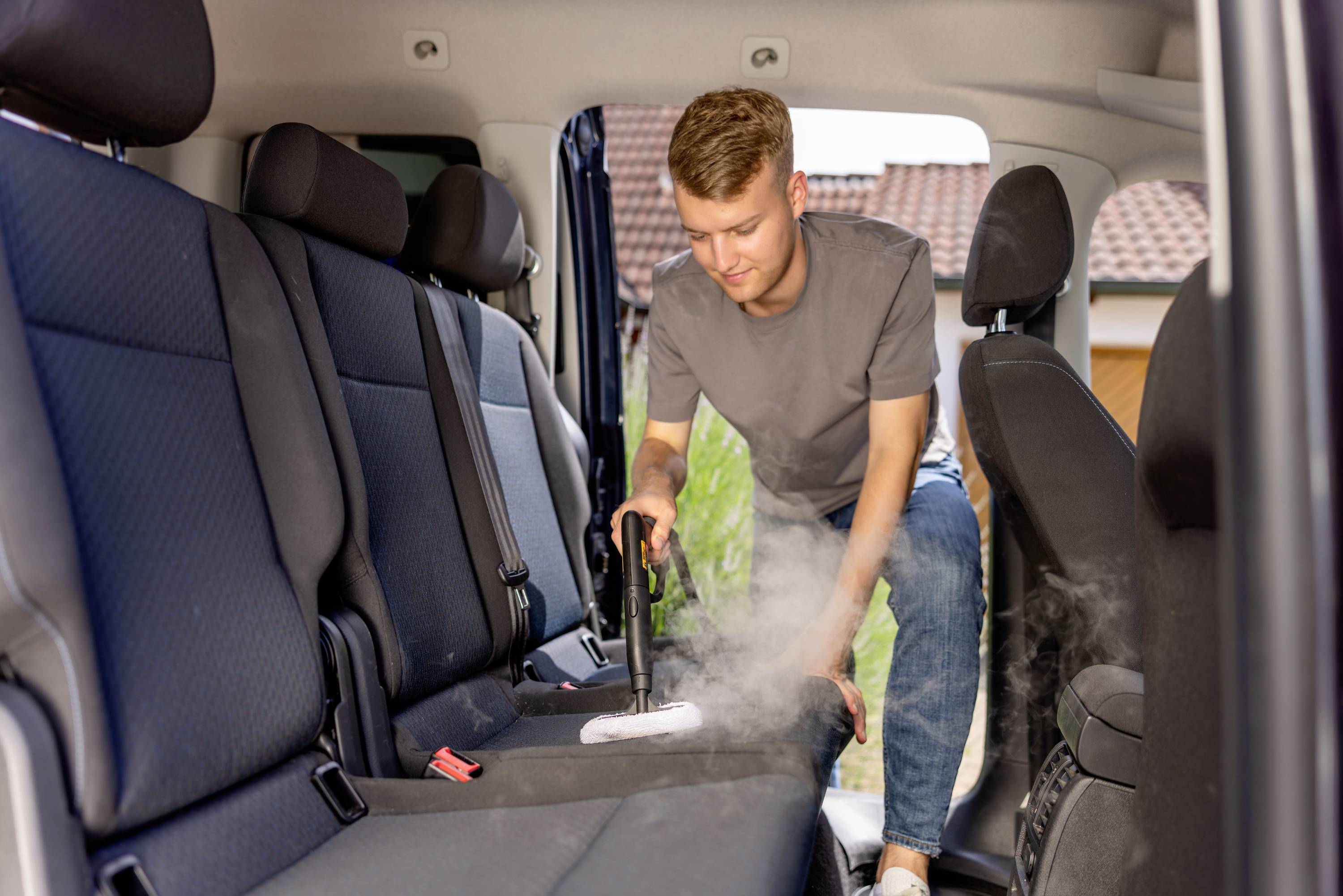 Mann reinigt Autoinnenraum mit Dampfreiniger. Er arbeitet an einem Sitz, trägt ein graues T-Shirt und blaue Jeans. Im Hintergrund sind Häuser sichtbar.