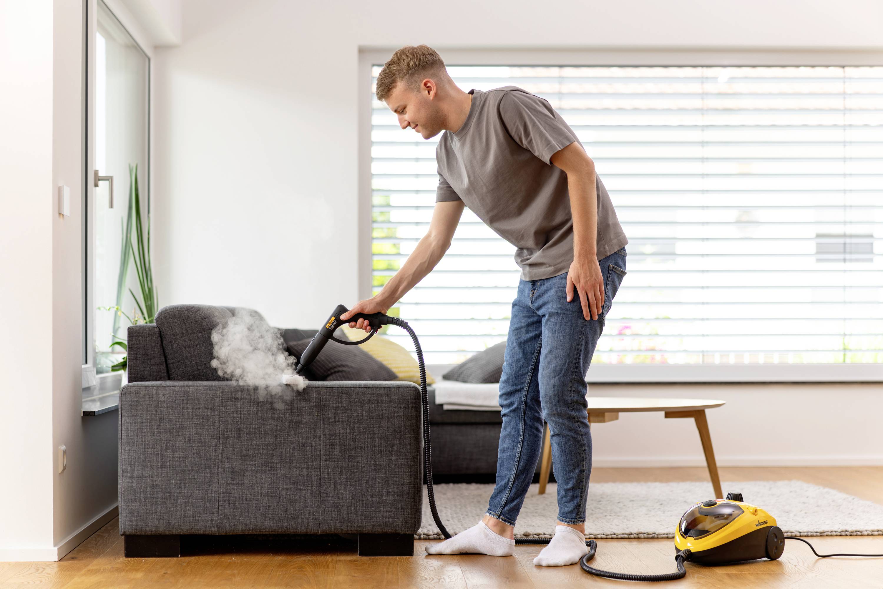 Ein Mann reinigt ein graues Sofa mit einem Dampfreiniger in einem hellen Wohnzimmer mit großen Fenstern im Hintergrund.