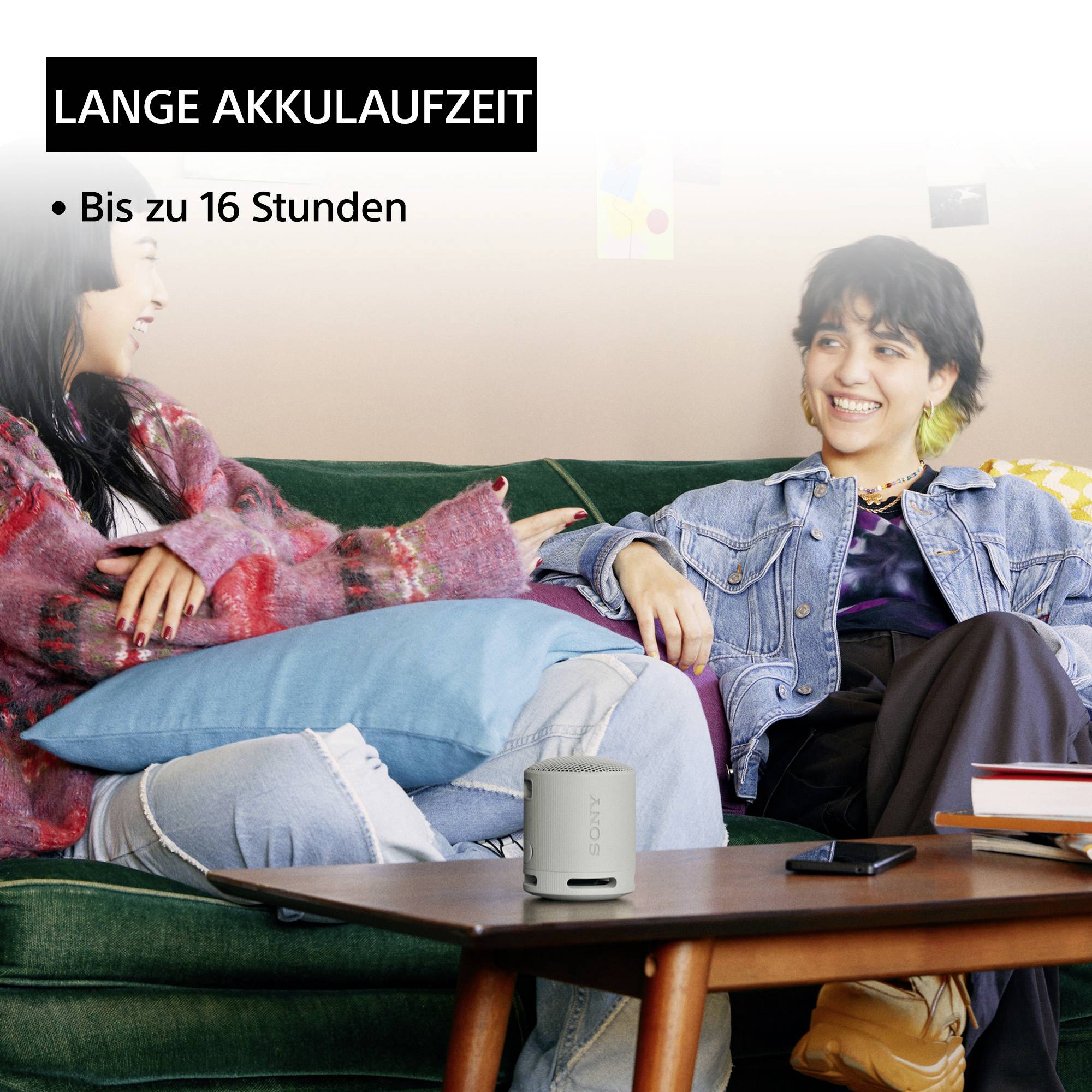 Zwei lachende Personen auf einem Sofa, die Musik aus einem tragbaren Lautsprecher hören. Text: 'Lange Akkulaufzeit bis zu 16 Stunden'.