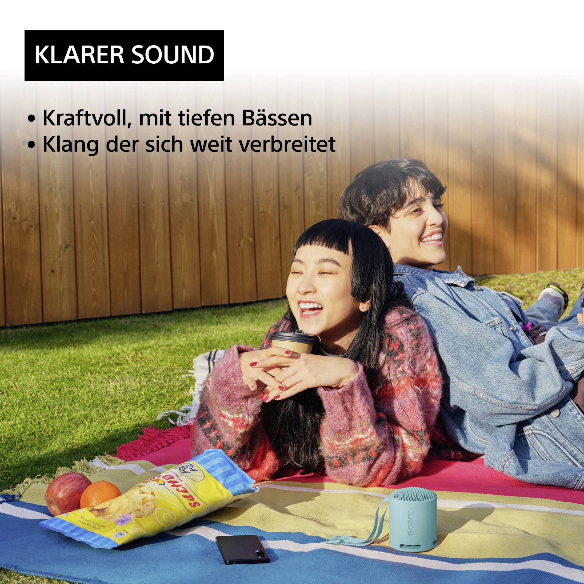 Zwei Personen sitzen auf einer Decke im Freien und hören Musik über einen tragbaren Lautsprecher. Neben ihnen liegen Chips und Obst.