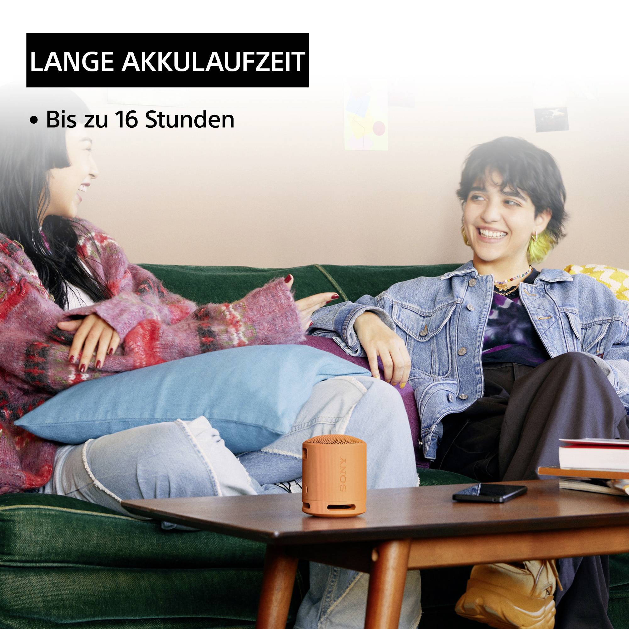 Zwei Personen lachen auf einem Sofa. Im Vordergrund liegt ein oranger Lautsprecher. Text oben links: 'Lange Akkulaufzeit, bis zu 16 Stunden'.