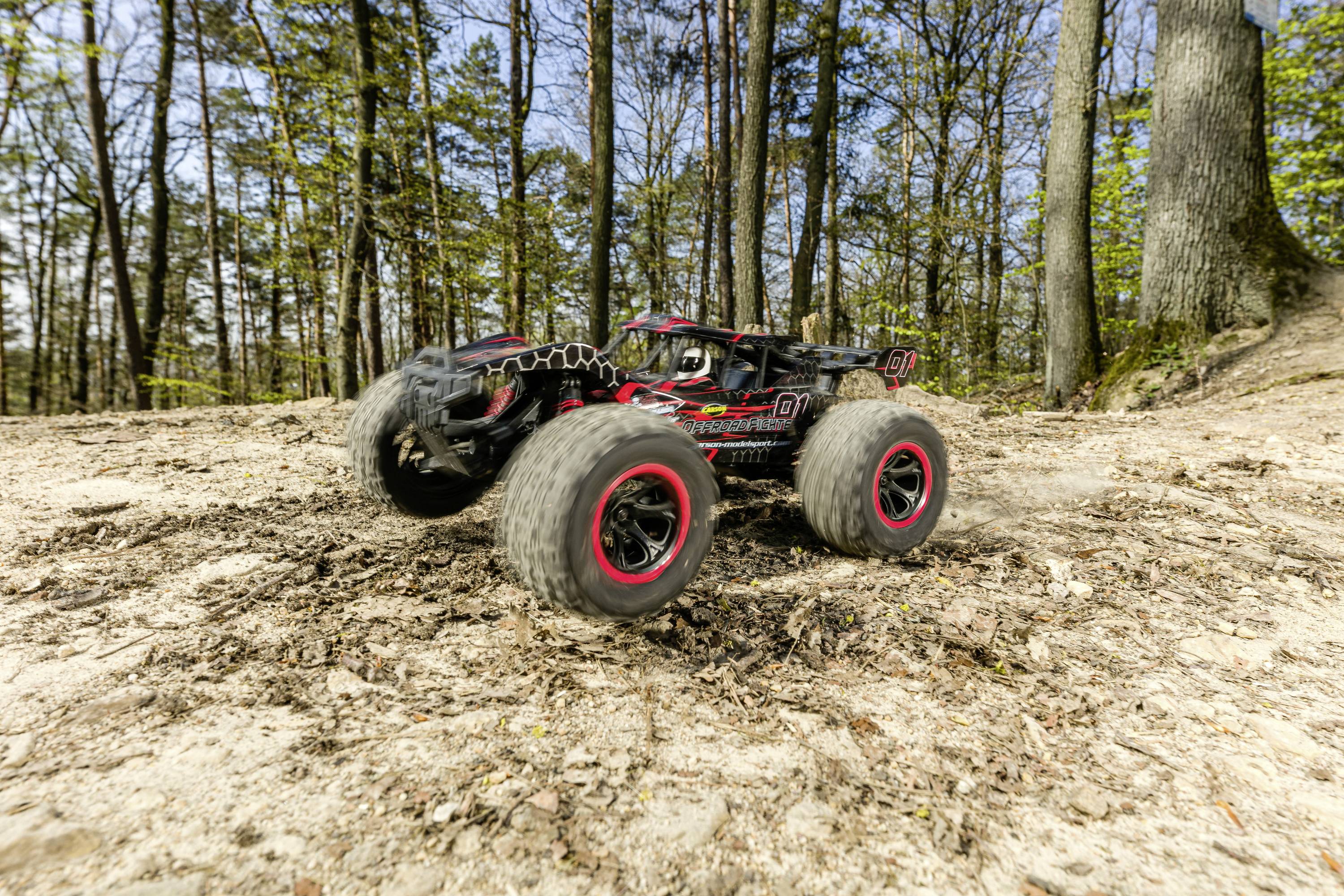 Ein ferngesteuertes Auto mit großen, roten Reifen fährt schnell durch einen Wald. Bäume und Laub im Hintergrund.