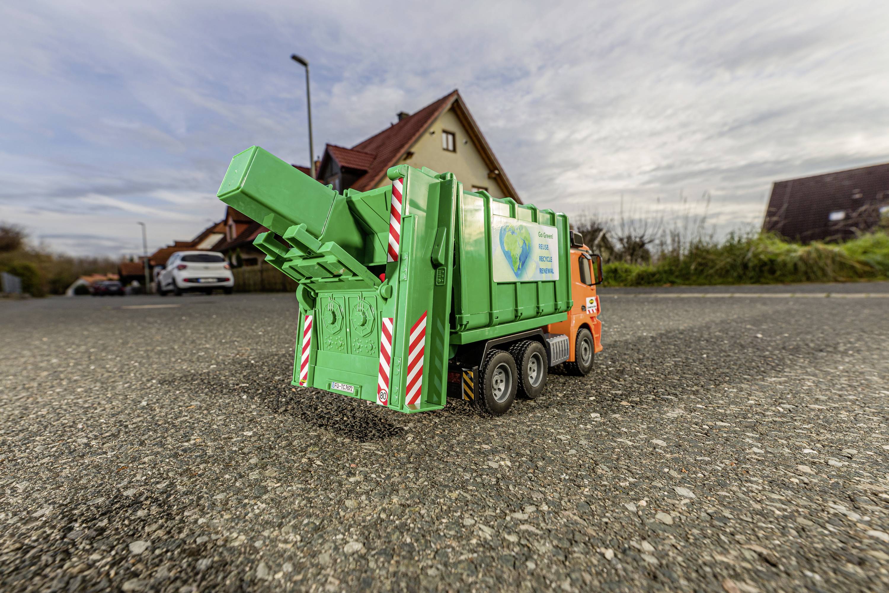 Ein orange-grüner Müllwagen steht auf einer Straße in einem Wohngebiet. Im Hintergrund sind Häuser und ein bewölkter Himmel zu sehen.