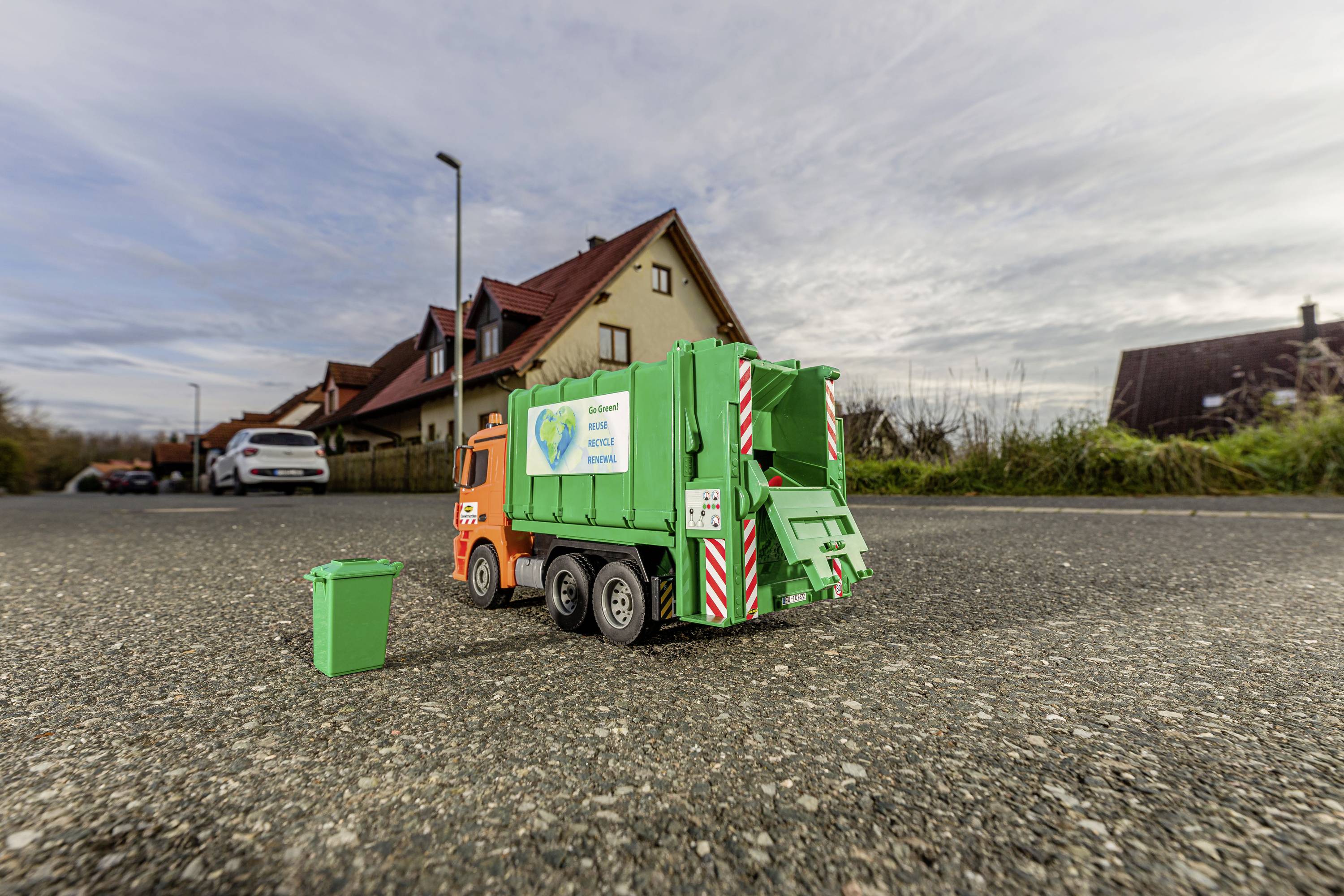 Ein kleines orange-grünes Spielzeug-Müllfahrzeug neben einem kleinen Mülleimer, auf einer asphaltierten Straße vor Häusern.