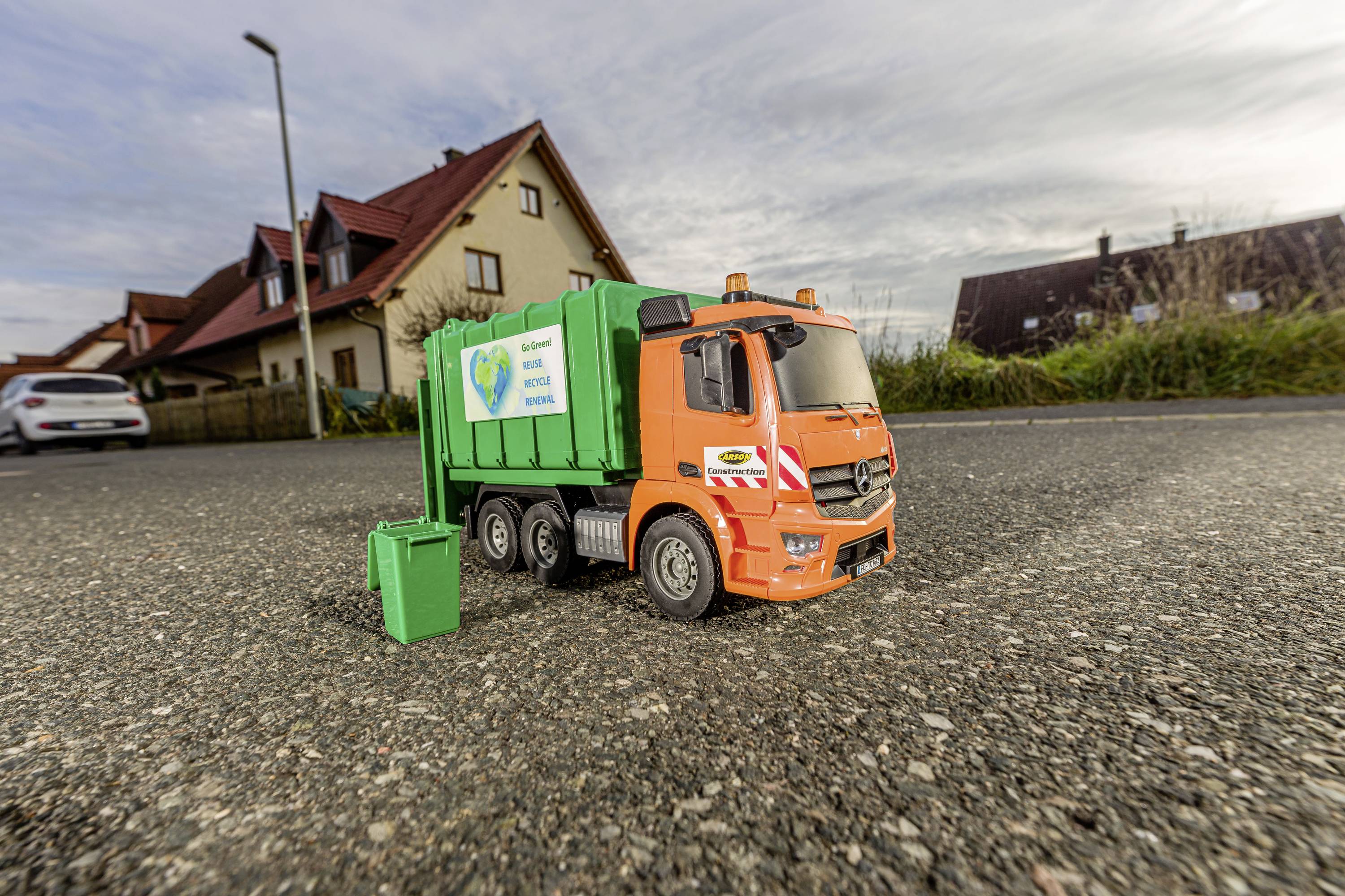 Spielzeug-Müllwagen auf einer Straße vor Häusern. Der orangefarbene LKW hat einen grünen Container mit Umwelt-Motiven.