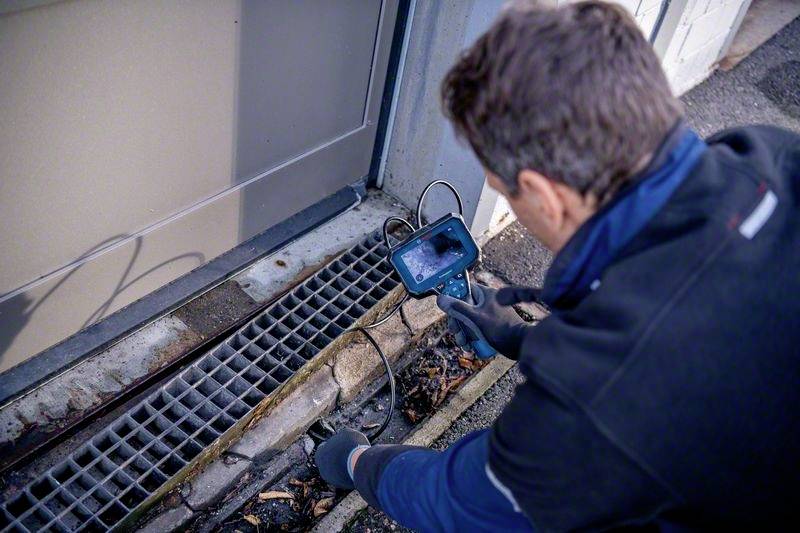 Ein Techniker überprüft mit einer Kamera eine Abflussrinne neben einem Gebäude auf Schäden oder Verstopfungen.