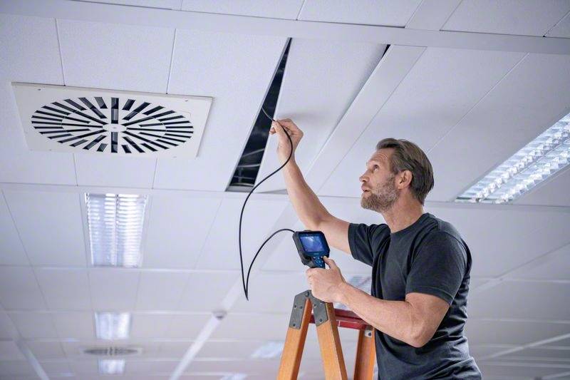 Ein Mann auf einer Leiter inspiziert eine Decke mit einer Kamera, die an einem Kabel befestigt ist, in einem Büro mit modernen Lichtern.