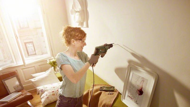 Eine Frau bohrt lächelnd ein Loch in eine Wand in einem hellen Wohnzimmer. Neben ihr liegt ein gerahmtes Bild auf einem Sofa.