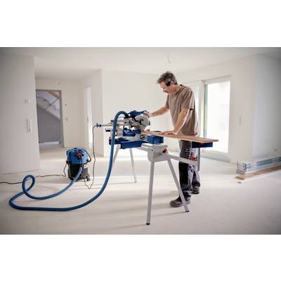 Ein Mann arbeitet in einem hellen Raum mit einer Gehrungssäge auf einem Tisch. Im Hintergrund sind weiße Wände und Fenster.