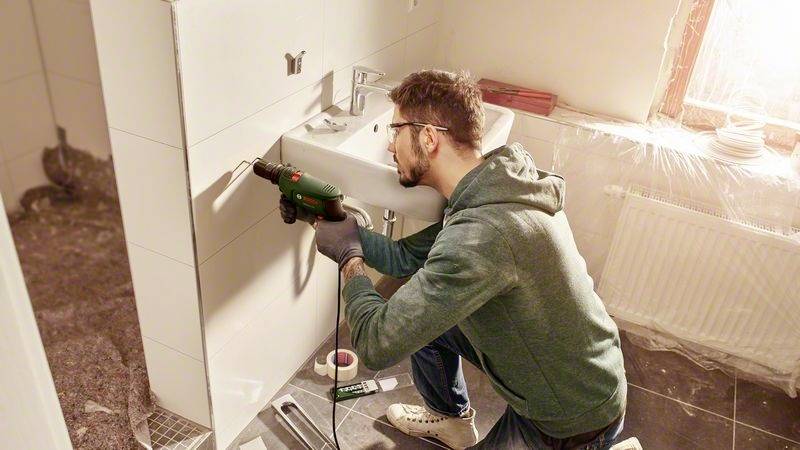 Ein Mann bohrt ein Loch in eine Badezimmerwand neben einem Waschbecken, möglicherweise zur Montage eines Badaccessoires.