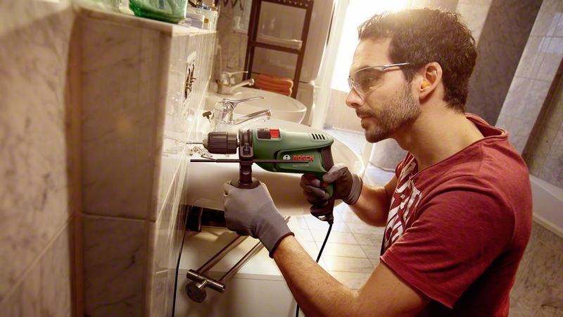 Ein Mann in Schutzbrille und Handschuhen bohrt mit einer Bohrmaschine in ein Waschbecken in einem Badezimmer.