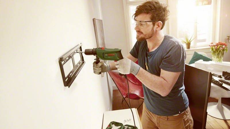 Ein Mann montiert eine TV-Halterung an eine Wand, während er eine Bohrmaschine benutzt. Er trägt Schutzbrille und Handschuhe, Tageslicht fällt durch das Fenster.