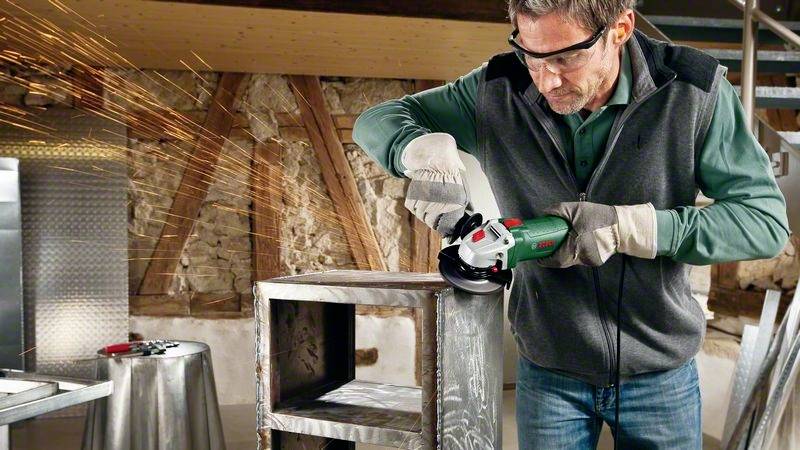 Ein Mann in Schutzkleidung schleift Metall mit einem Winkelschleifer in einer Werkstatt. Funken fliegen vom Metall ab.
