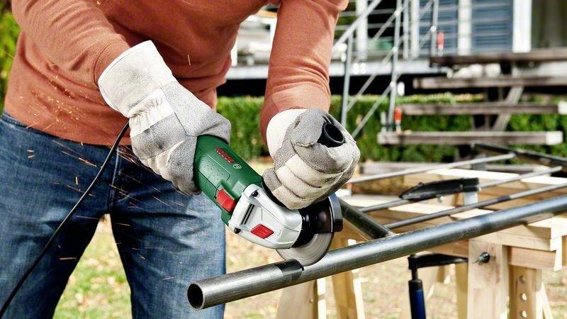 Person mit Schutzkleidung verwendet eine elektrische Schleifmaschine, um ein Metallrohr auf einer Arbeitsfläche im Freien zu bearbeiten.