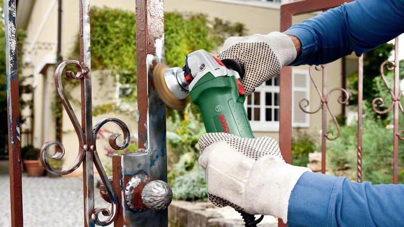 Eine Person schleift mit einem Winkelschleifer Rost von einem Metalltor ab. Sie trägt blaue Kleidung und Arbeitshandschuhe.