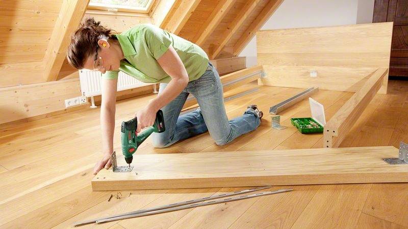 Eine Frau montiert ein Holzregal im Dachboden. Sie trägt Gehörschutz und nutzt einen Akkuschrauber. Um sie herum liegen Werkzeug und Bretter.