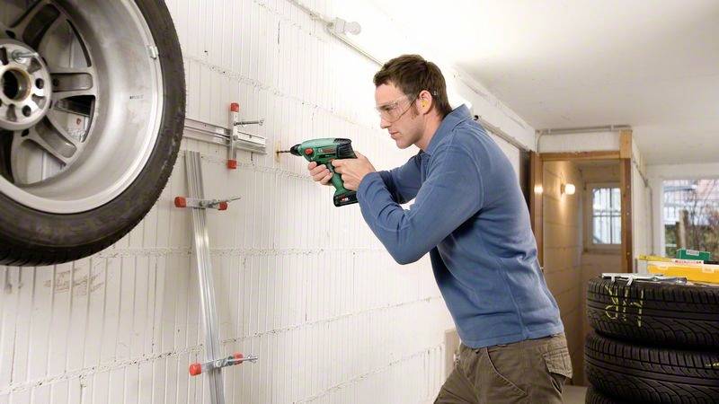 Eine Person montiert ein Ersatzreifenregal an eine Wand in einer Garage, während sie eine Schutzbrille trägt.