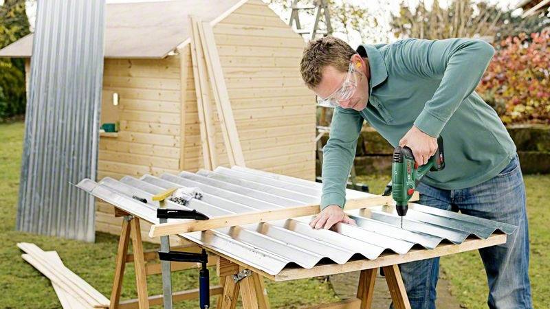 Ein Mann in Schutzbrille schraubt Wellblech auf einem Arbeitstisch im Garten. Im Hintergrund ist ein Holzschuppen zu sehen.