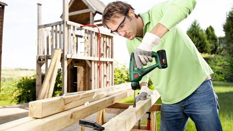 Mann mit Schutzbrille und Handschuhen schraubt im freien Gartenbereich Holzlatten zusammen. Im Hintergrund Spielplatz.