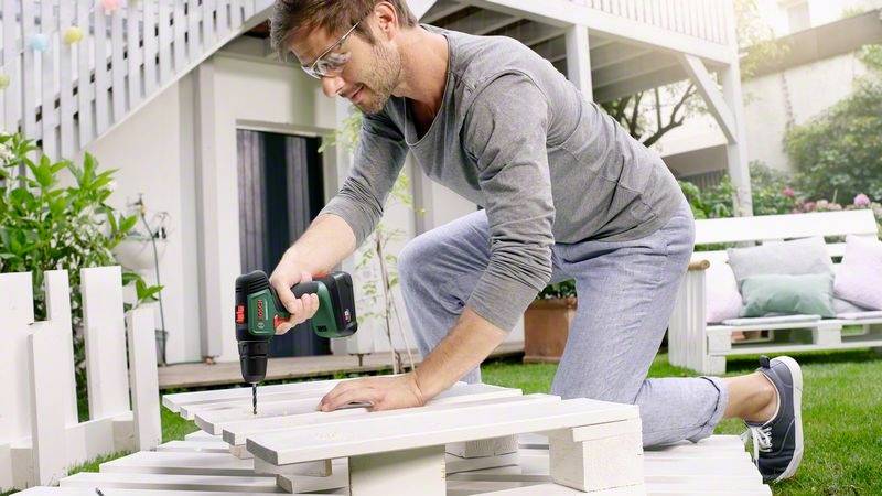 Ein Mann kniet im Garten und schraubt mit einem Akkuschrauber Holzbretter zusammen, wahrscheinlich für ein Möbelstück im Freien.