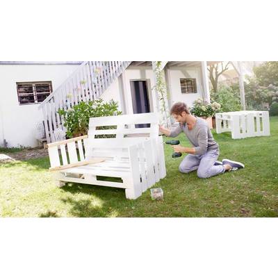 Eine Person baut im Garten eine weiße Holzbank zusammen. Im Hintergrund ist ein Haus mit Terrasse sichtbar.